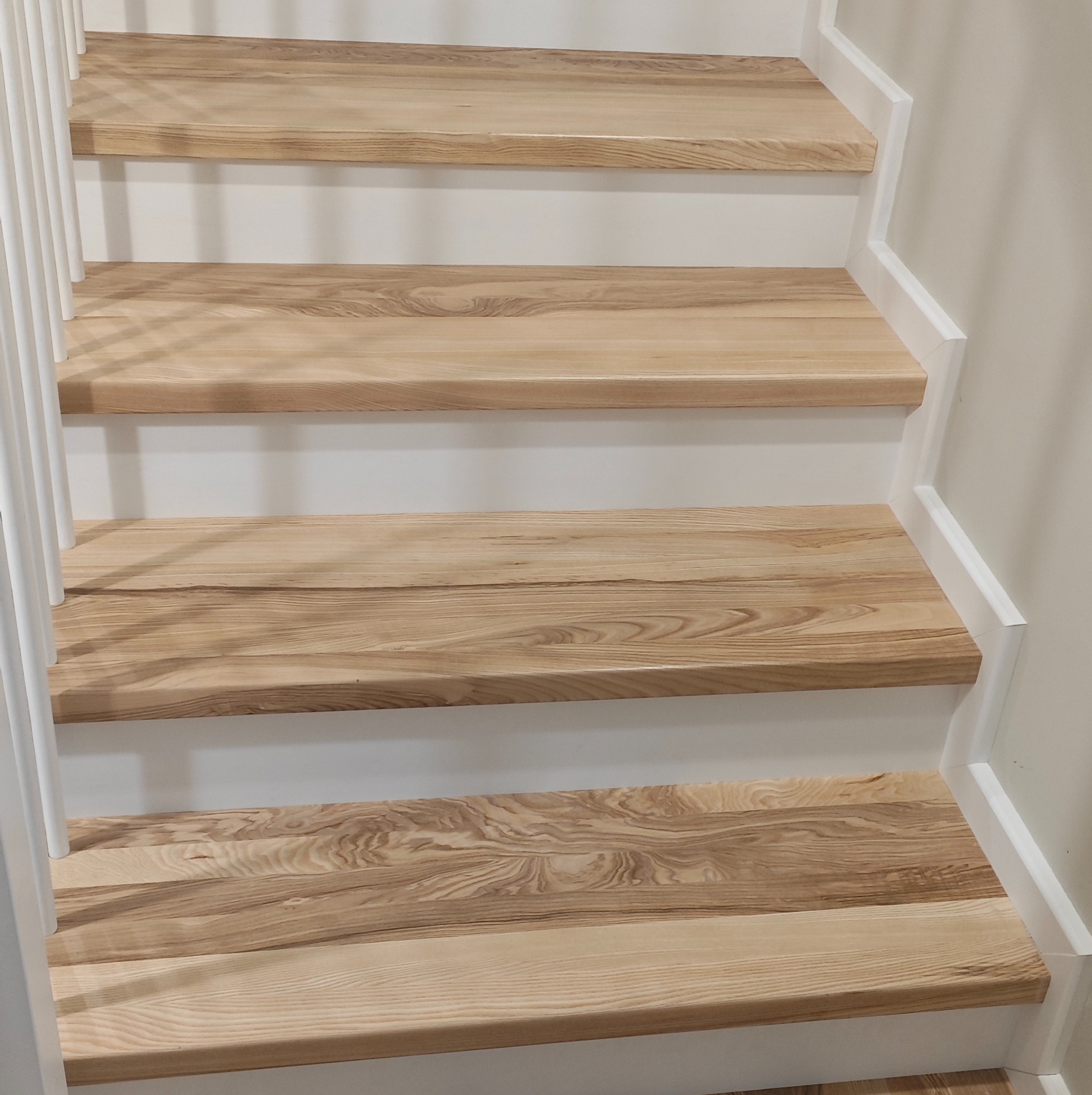 Jasne, drewniane schody z białymi podstopnicami i balustradą. Widoczne usłojenie drewna. Kąt ujęcia od dołu, skupiony na detalach stopni.