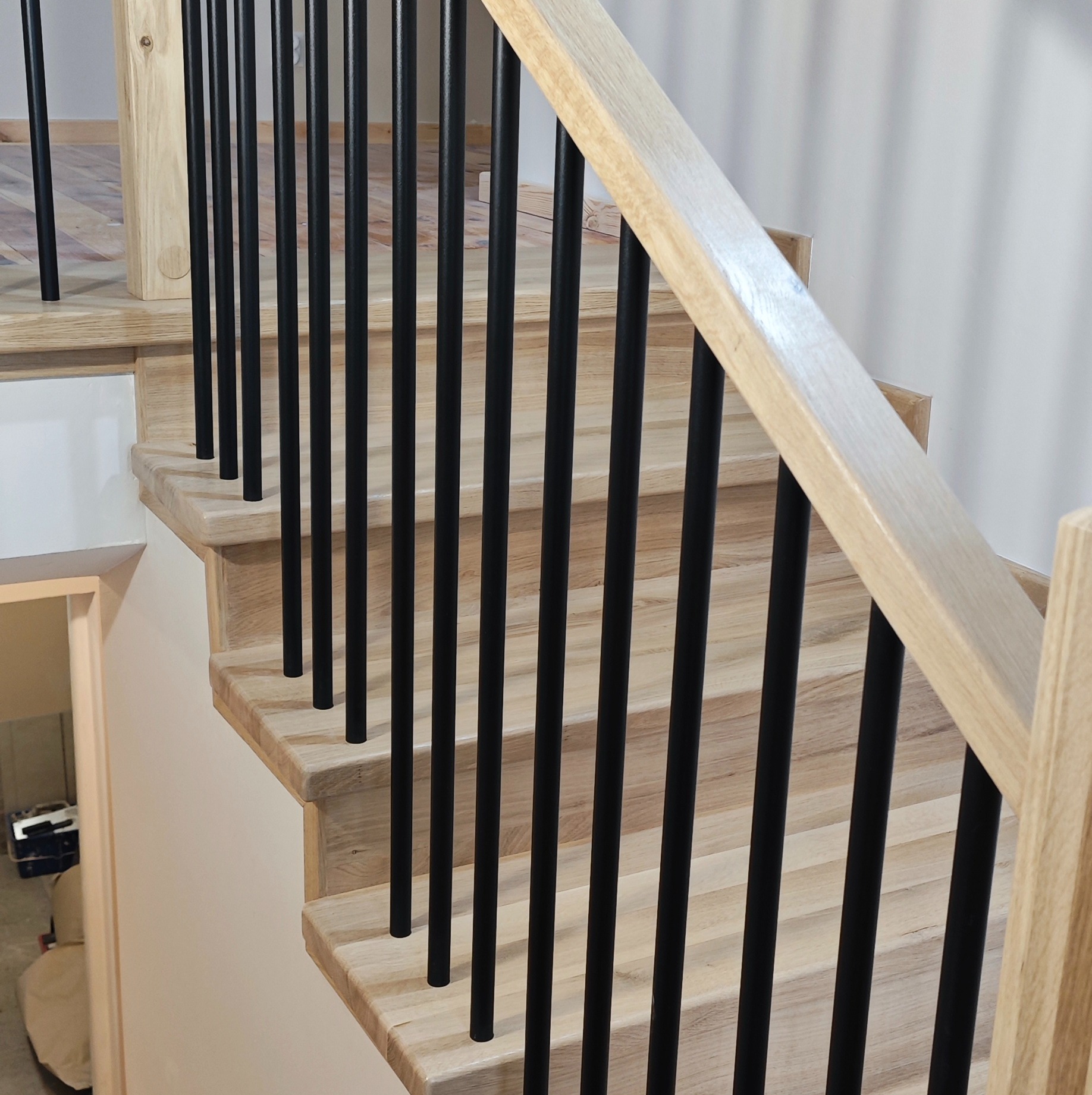 Jasne drewniane schody z czarnymi, metalowymi balustradami w nowoczesnym wnętrzu. Widoczne detale wykończenia i naturalne usłojenie drewna.