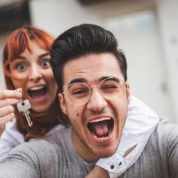 Ekstatyczna para, on w okularach z otwartymi ustami i przymrużonym okiem, ona z kluczami w dłoni, świętuje zakup mieszkania na tle jasnej elewacji budynku.