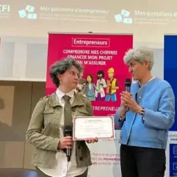 Wręczenie nagrody na konferencji, kobieta z dyplomem i mikrofonem, obok niej kobieta w niebieskiej marynarce, w tle banery z informacjami o finansach i przedsiębiorczości.