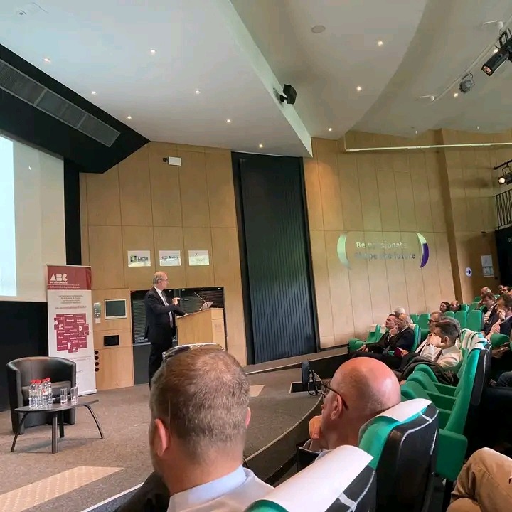 Prelegent w garniturze wygłasza prezentację podczas seminarium dla grupy osób siedzących na zielonych krzesłach w audytorium.