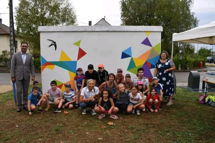 Grupa dzieci i dorosłych pozuje przed pomalowaną na biało skrzynką transformatorową z geometrycznym wzorem i sylwetką jaskółki, na trawniku w plenerze.