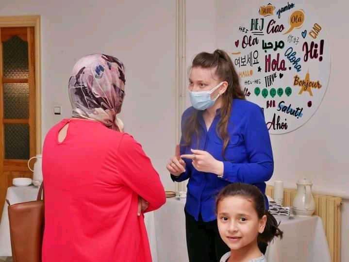 Kobieta w niebieskiej bluzce i maseczce gestykuluje, rozmawiając z kobietą w różowej sukience i hidżabie, w tle dziecko i okrągła tablica z powitaniami w różnych językach.