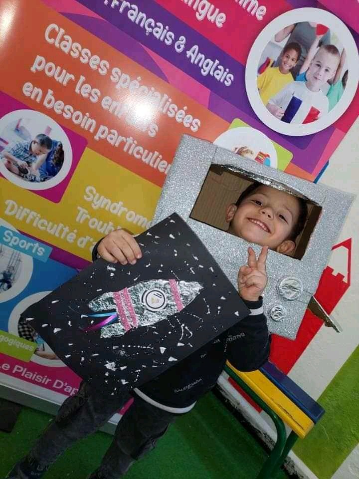 Uśmiechnięty chłopiec w Polonisz przebrany za telewizor, prezentuje czarną kartkę z narysowaną rakietą, na tle plakatu z informacjami o zajęciach specjalistycznych.