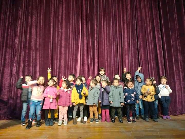 Grupa dzieci w różnym wieku stoi na scenie przed bordową kurtyną, pozując i gestykulując radośnie.