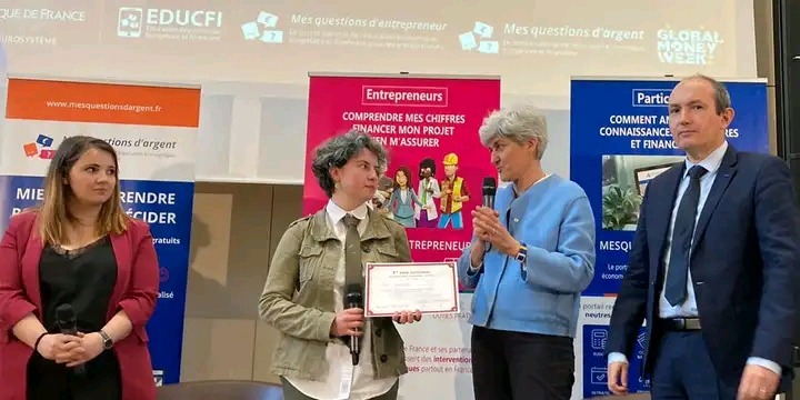 Wręczenie nagrody na konferencji, kobieta z dyplomem i mikrofonem, obok niej kobieta w niebieskiej marynarce, w tle banery z informacjami o finansach i przedsiębiorczości.