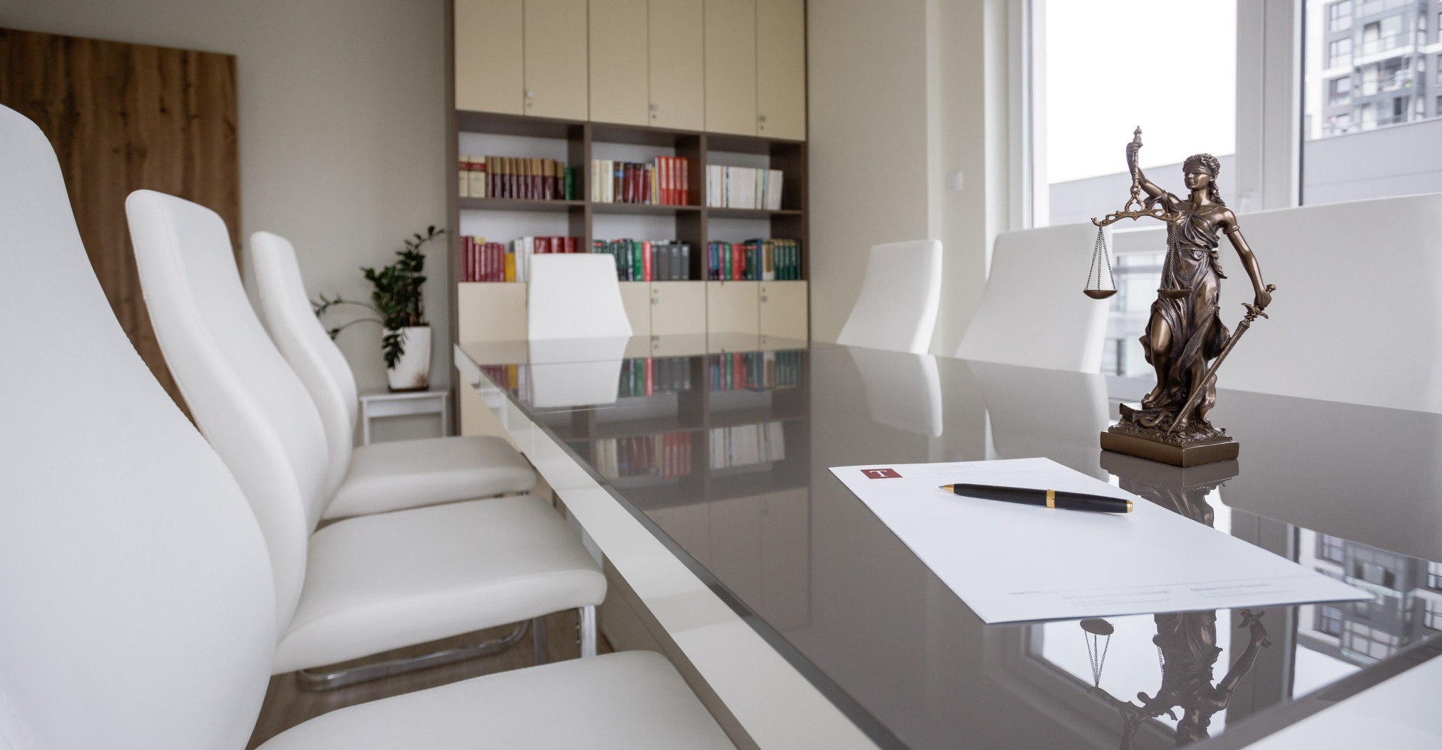 Sala konferencyjna w kancelarii prawnej z długim szarym stołem, białymi krzesłami, regałem z książkami w tle oraz figurką Temidy, dokumentem i piórem na blacie.