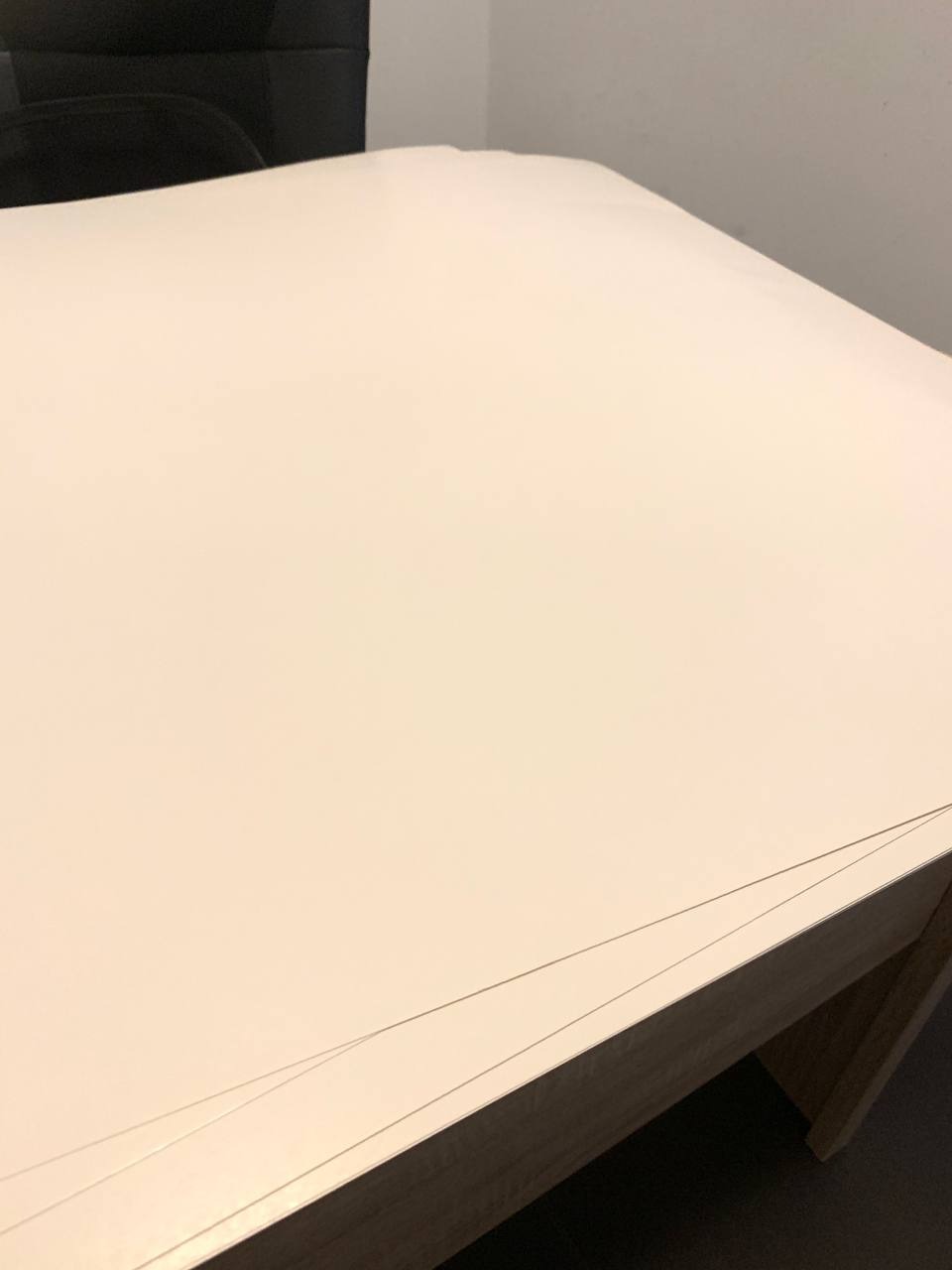 Stos arkuszy białego, gładkiego papieru na drewnianym podłożu, widoczne krawędzie arkuszy i fragment czarnego krzesła w tle.