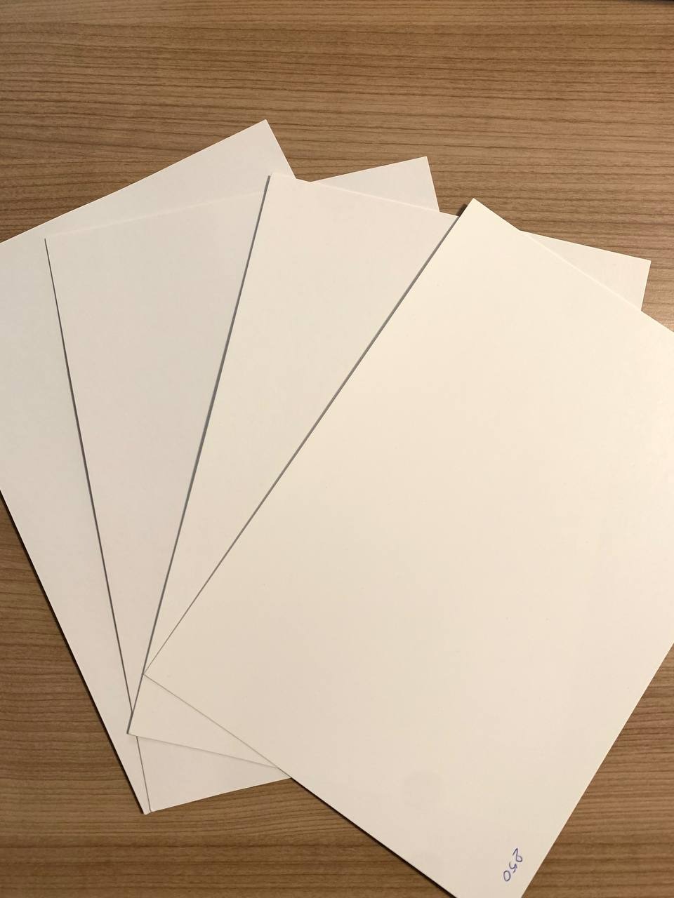 Stos białych arkuszy papieru ułożonych ukośnie na drewnianym blacie, z widocznym odręcznym napisem '250' w prawym dolnym rogu.