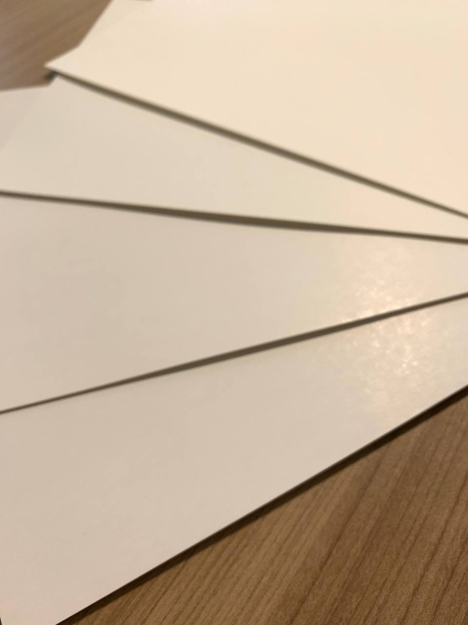 Stos arkuszy białego, gładkiego papieru leżących na drewnianej powierzchni, z widocznymi cieniami krawędzi.