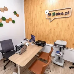 Nowoczesne biuro dietetyka z drewnianymi panelami na ścianie, dekoracjami z mchu w kształcie plastrów miodu, biurkiem z laptopem i sprzętem medycznym na kółkach.