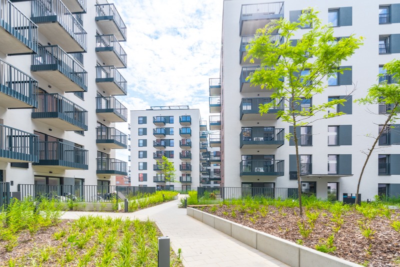 Nowoczesne osiedle mieszkaniowe z białymi budynkami, ciemnymi balkonami i zadbaną zielenią, widok z poziomu chodnika.