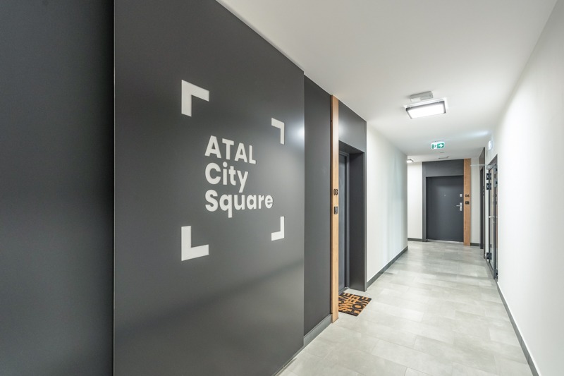 Wnętrze korytarza budynku mieszkalnego z ciemnoszarą ścianą z logo 'ATAL City Square' otoczonego białymi kwadratowymi nawiasami, widoczne drzwi do windy z numerem '03', wycieraczka i drzwi...