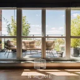 Widok z wnętrza przez duże okno balkonowe na taras z rattanowymi fotelami i roślinami w donicach, w tle błękitne niebo.