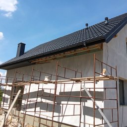 Dom jednorodzinny w trakcie ocieplania elewacji, widoczne rusztowania i fragment czarnego dachu z kominem na tle błękitnego nieba.