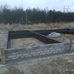 Wzniesione mury fundamentowe z bloczków betonowych, zabezpieczone czarną izolacją, na tle leśnego krajobrazu i pochmurnego nieba. Widoczne drewniane stemple stabilizujące.