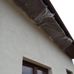 Fragment elewacji domu z oknami i zabezpieczonym okapem folią malarską. Świeżo nałożony tynk strukturalny w jasnym odcieniu. Ujęcie z dołu, podkreślające fakturę.