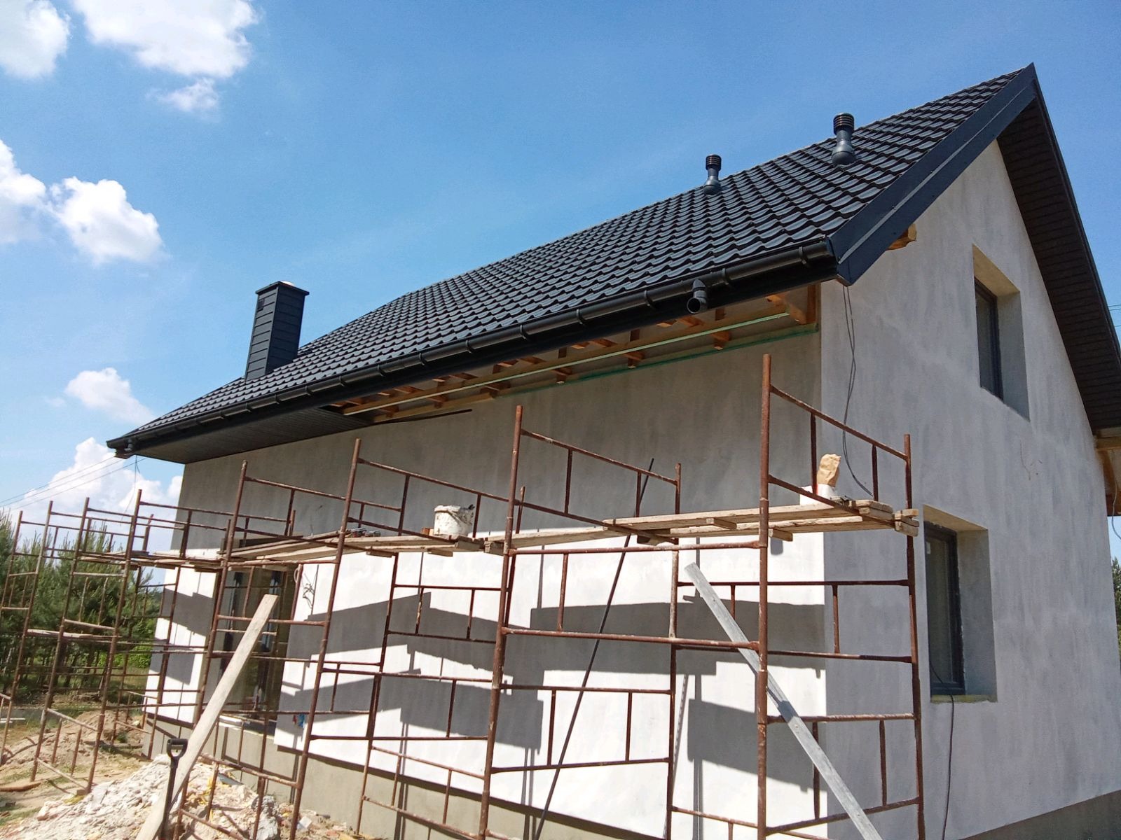 Dom jednorodzinny w trakcie ocieplania elewacji, widoczne rusztowania i fragment czarnego dachu z kominem na tle błękitnego nieba.