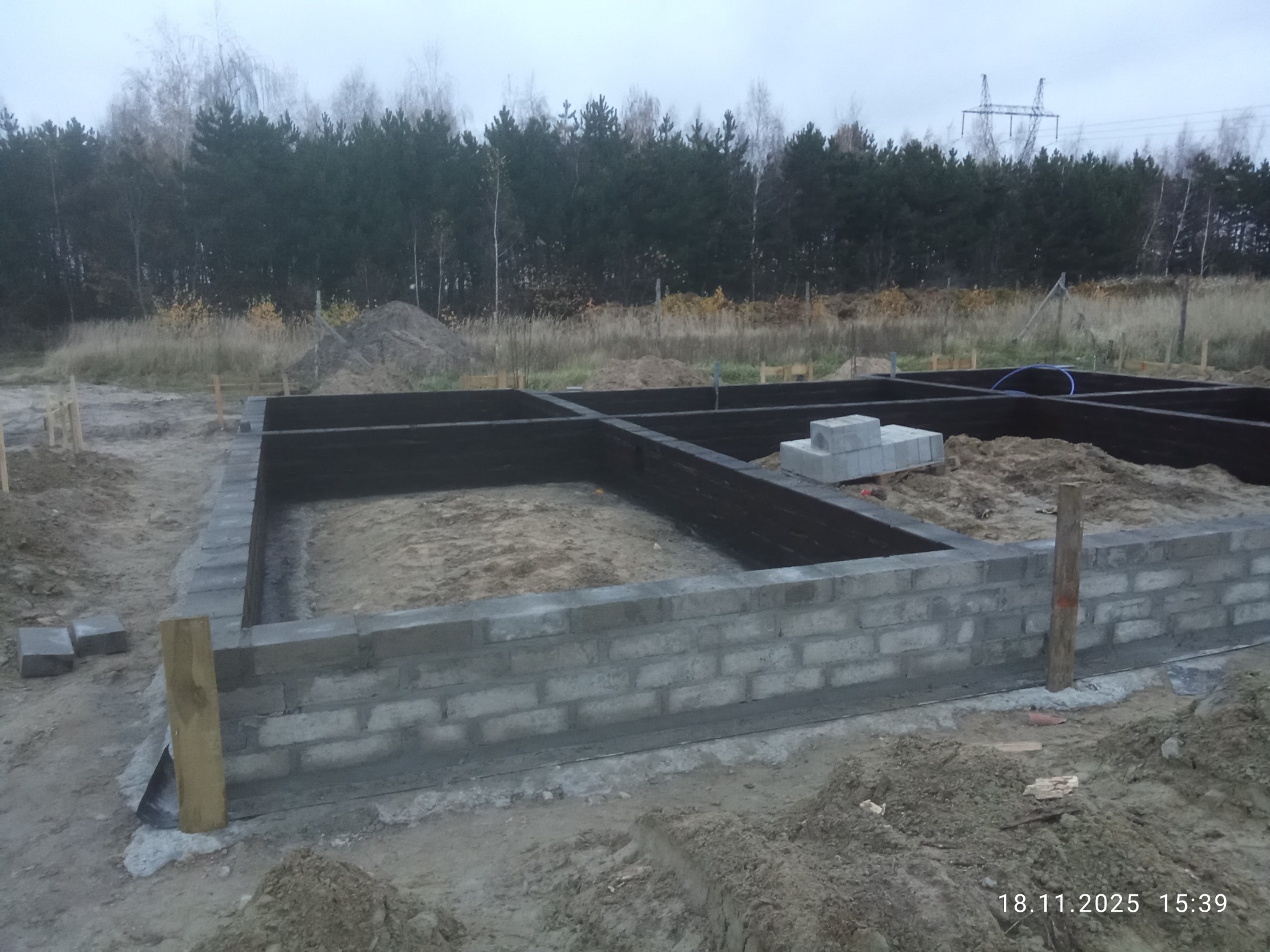 Wzniesione mury fundamentowe z bloczków betonowych, zabezpieczone czarną izolacją, na tle leśnego krajobrazu i pochmurnego nieba. Widoczne drewniane stemple stabilizujące.