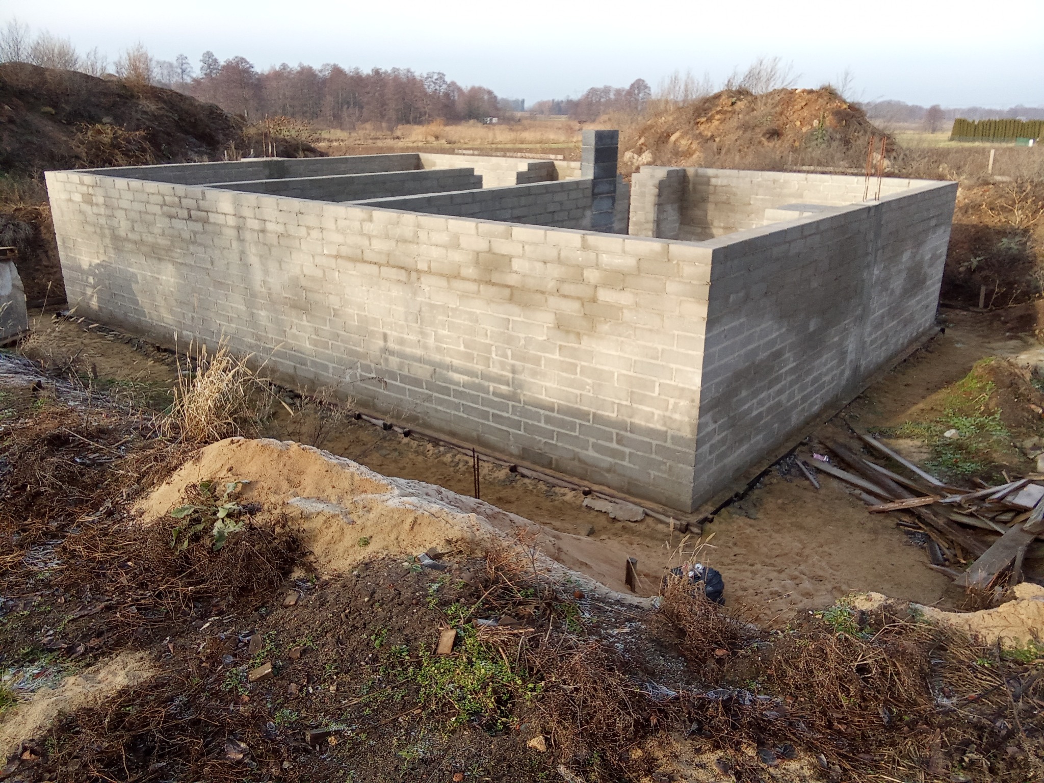 Fundamenty domu z bloczków betonowych na działce, widok z góry. Częściowo wykopane, zbrojenie wystaje. W tle drzewa i ziemia.