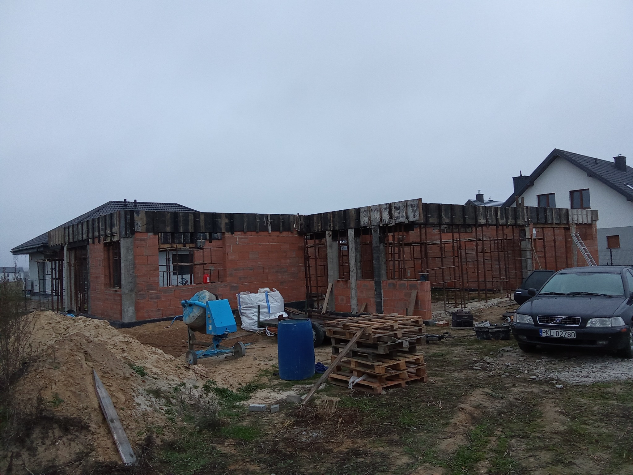 Surowy stan domu z czerwonej cegły w trakcie budowy, widoczna betoniarka, palety i samochód osobowy na tle pochmurnego nieba. W tle dom sąsiada.