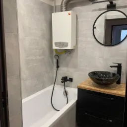 Nowoczesna łazienka z szarymi płytkami, białą wanną, czarną armaturą i okrągłym lustrem; na ścianie biały piec gazowy z żółtą rurą.