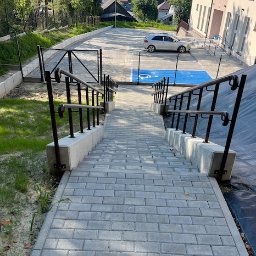 STimpill - Przedsiębiorstwo Budowlane - Schody z kostki brukowej prowadzące na parking z wyznaczonym miejscem dla niepełnosprawnych, metalowe barierki po bokach, budynek w tle, ujęcie z góry.