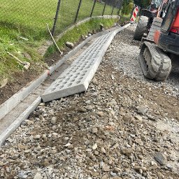 STimpill - Przedsiębiorstwo Budowlane - Wykonywanie korytowania pod drogę w Mucharzu, z widoczną betonową rynną i ażurowymi płytami oraz fragmentem koparki gąsienicowej.