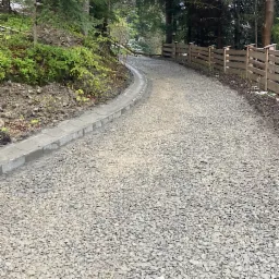 Żwirowa droga dojazdowa w lesie, z betonowym korytkiem odwadniającym po lewej stronie i drewnianym ogrodzeniem po prawej, widok z poziomu gruntu.