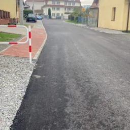 Świeżo położony, czarny asfalt na drodze w małej miejscowości, widok na budynek z wieżą w tle.