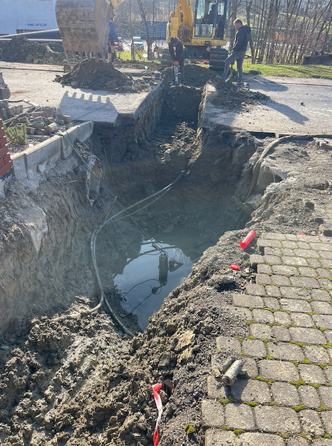 Głęboki wykop pod instalacje, częściowo zalany wodą, z widocznymi kablami i rurami, wykonywany przy użyciu koparki i młota pneumatycznego, w tle budynek i drzewa.