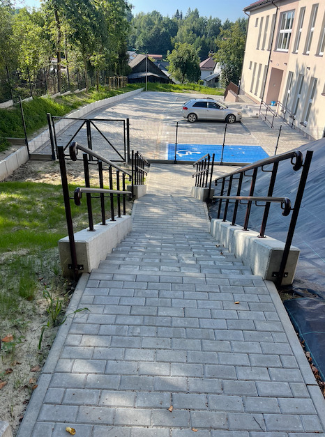 Schody z kostki brukowej prowadzące na parking z wyznaczonym miejscem dla niepełnosprawnych, metalowe barierki po bokach, budynek w tle, ujęcie z góry.