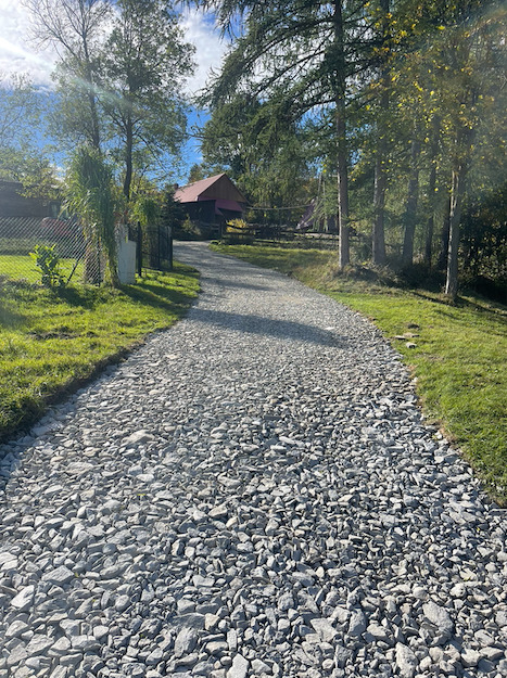 Ujęcie nowo wysypanej, szarej drogi z kamienia łamanego, prowadzącej w kierunku zabudowań gospodarczych z czerwonym dachem, otoczonej zielenią drzew i trawników.