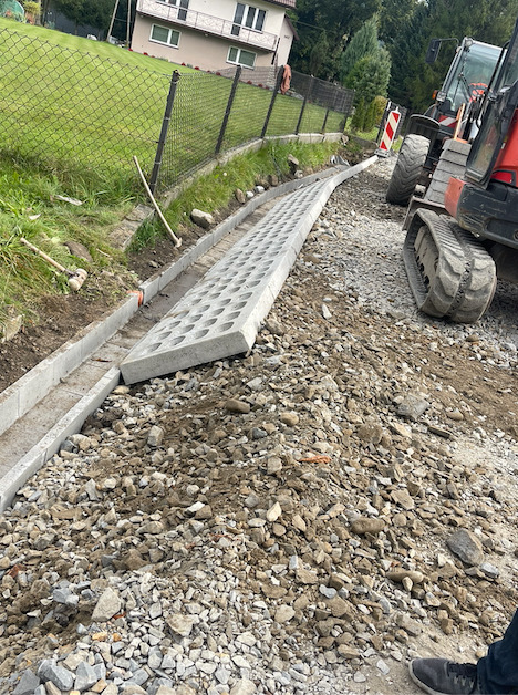 Wykonywanie korytowania pod drogę w Mucharzu, z widoczną betonową rynną i ażurowymi płytami oraz fragmentem koparki gąsienicowej.