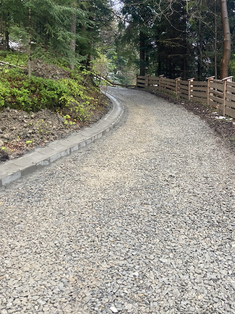 Żwirowa droga dojazdowa w lesie, z betonowym korytkiem odwadniającym po lewej stronie i drewnianym ogrodzeniem po prawej, widok z poziomu gruntu.