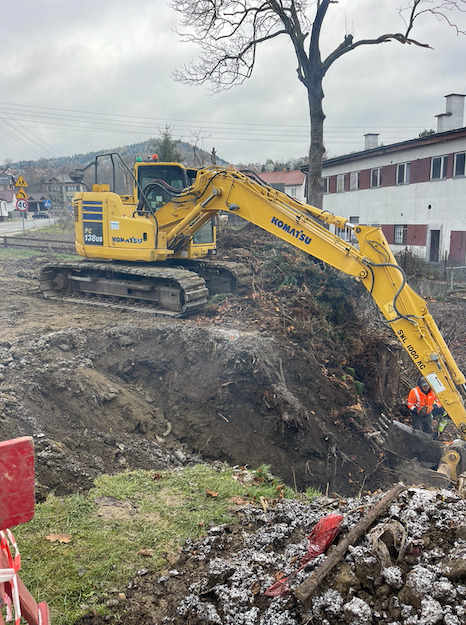 Żółta koparka Komatsu PC138US w trakcie prac ziemnych, widoczny wykop w gruncie, w tle budynek i drzewa, pochmurne niebo.
