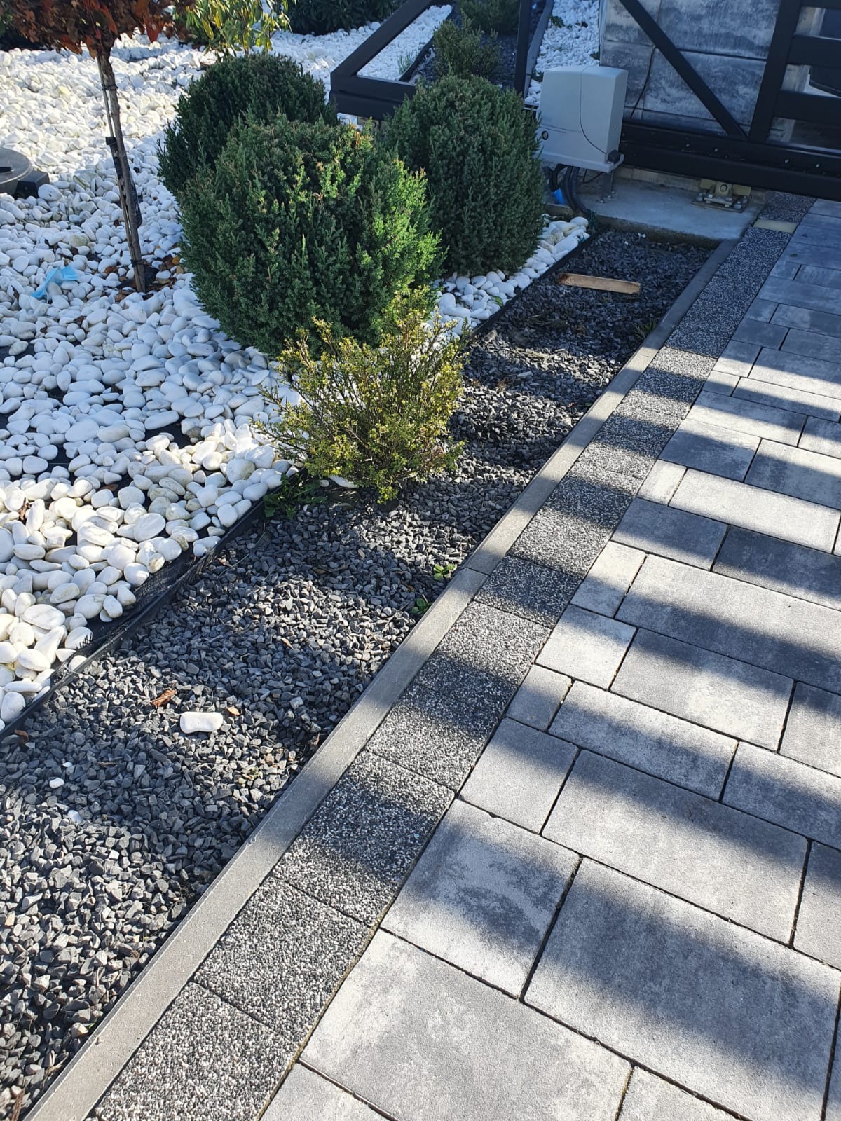 Wykończony fragment ogrodu z ozdobnymi kamieniami (białe i szare) oraz niskimi krzewami iglastymi i chodnikiem z szarych płyt, widok z góry.