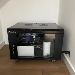Czarna szafa rack Lanberg z przeszklonymi drzwiami, zawierająca router Huawei, router TP-Link, przełącznik sieciowy i inne urządzenia, umieszczona w rogu pomieszczenia z drewnianą podłogą.