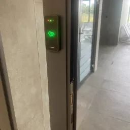 Zewnętrzny czytnik kart zbliżeniowych Roger z zieloną ikoną karty na szarej ramie drzwiowej, widok z boku na przeszklone drzwi i fragment tarasu.