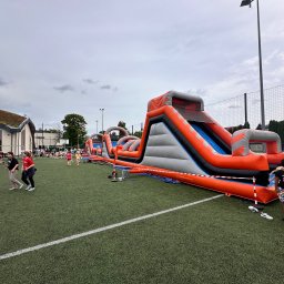 Fun Factory - Dmuchany tor przeszkód z wysoką zjeżdżalnią na zielonym boisku. W tle grupa dzieci i dorosłych, oświetlenie boiskowe. Pomarańczowo-szara konstrukcja.