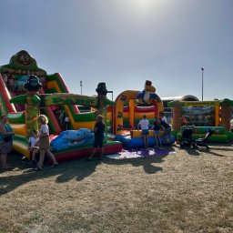 Fun Factory - Kolorowe dmuchańce z motywem dżungli, w tym zjeżdżalnia i mały basen, ustawione na trawie, z bawiącymi się dziećmi i dorosłymi w słoneczny dzień.