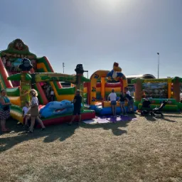 Kolorowe dmuchańce z motywem dżungli, w tym zjeżdżalnia i mały basen, ustawione na trawie, z bawiącymi się dziećmi i dorosłymi w słoneczny dzień.