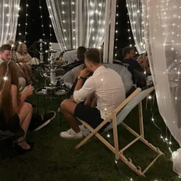Grupa osób relaksuje się w namiocie oświetlonym girlandami, z widocznymi fajkami wodnymi i drinkami na stolikach.