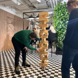 Grupa osób gra w drewnianą wieżę na czarno-białej podłodze w biurze z zieloną ścianą, widoczny mężczyzna w zielonej bluzie wyciągający klocek.