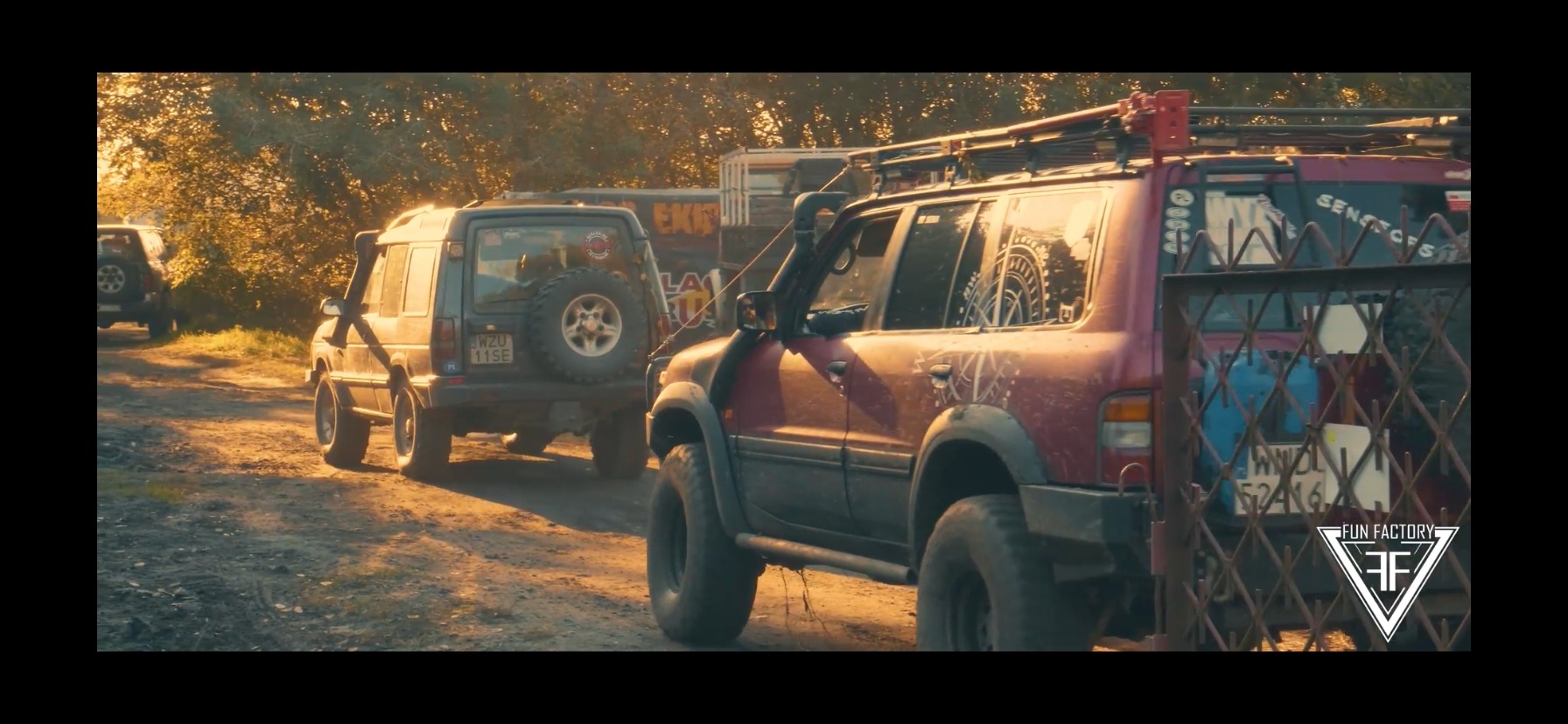 Dwa samochody terenowe przygotowane do jazdy off-road, z widocznymi modyfikacjami i zabrudzeniami po jeździe w terenie, ustawione na polnej drodze w słoneczny dzień.