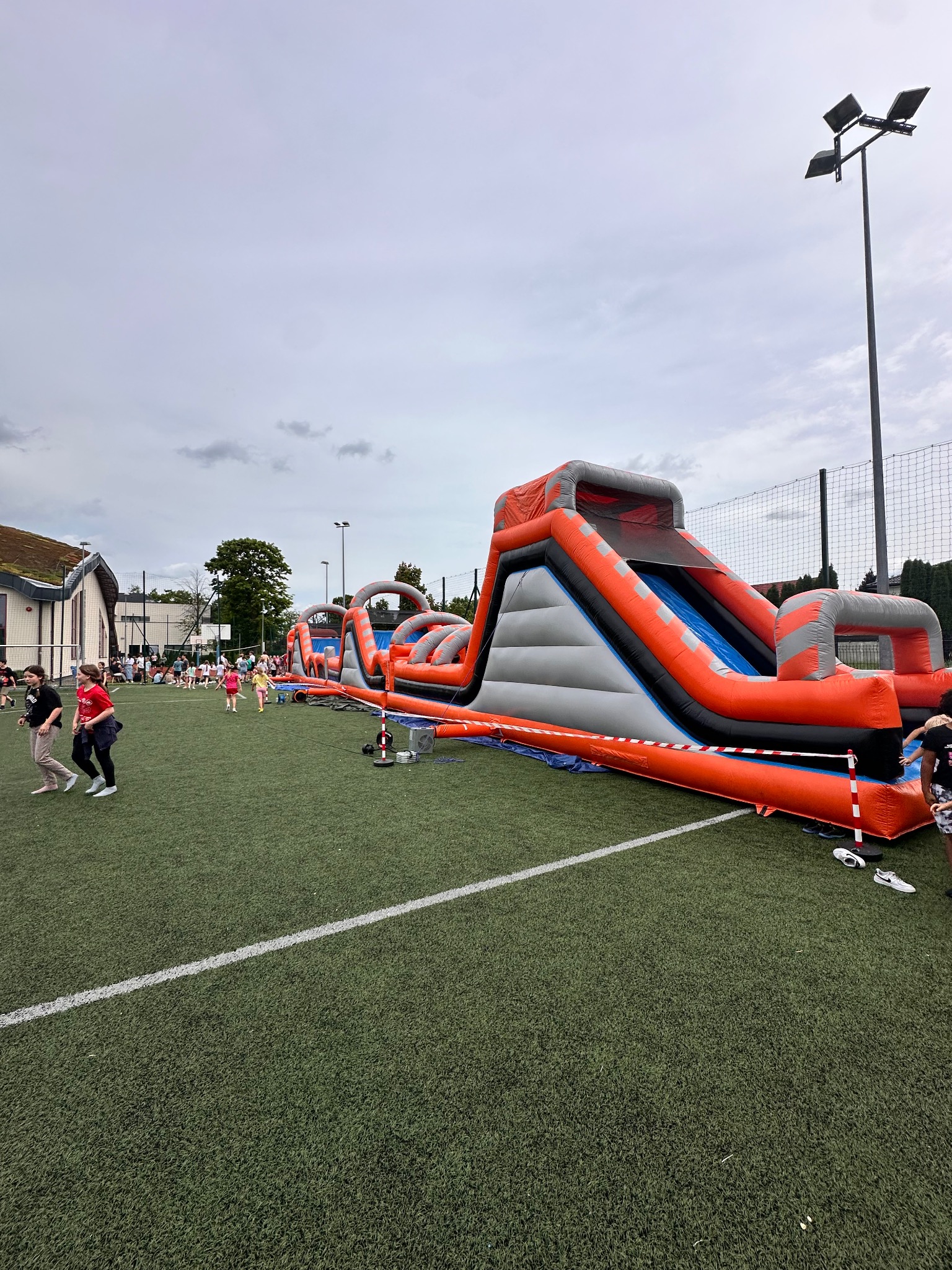 Dmuchany tor przeszkód z wysoką zjeżdżalnią na zielonym boisku. W tle grupa dzieci i dorosłych, oświetlenie boiskowe. Pomarańczowo-szara konstrukcja.