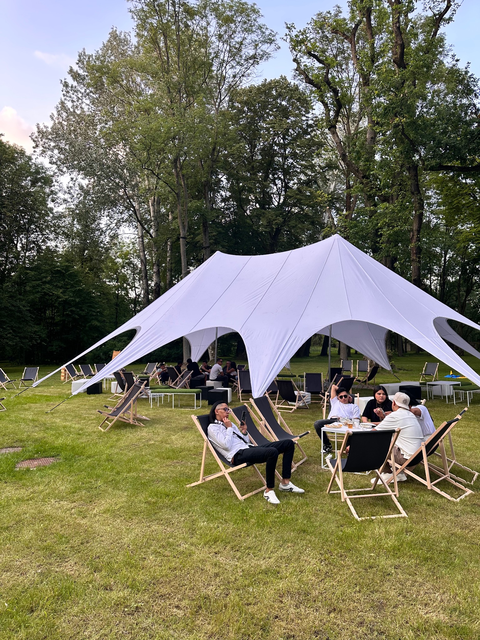 Biały namiot gwiazda na zielonej trawie, otoczony leżakami i stolikami z ludźmi. Plenerowa impreza w otoczeniu drzew.