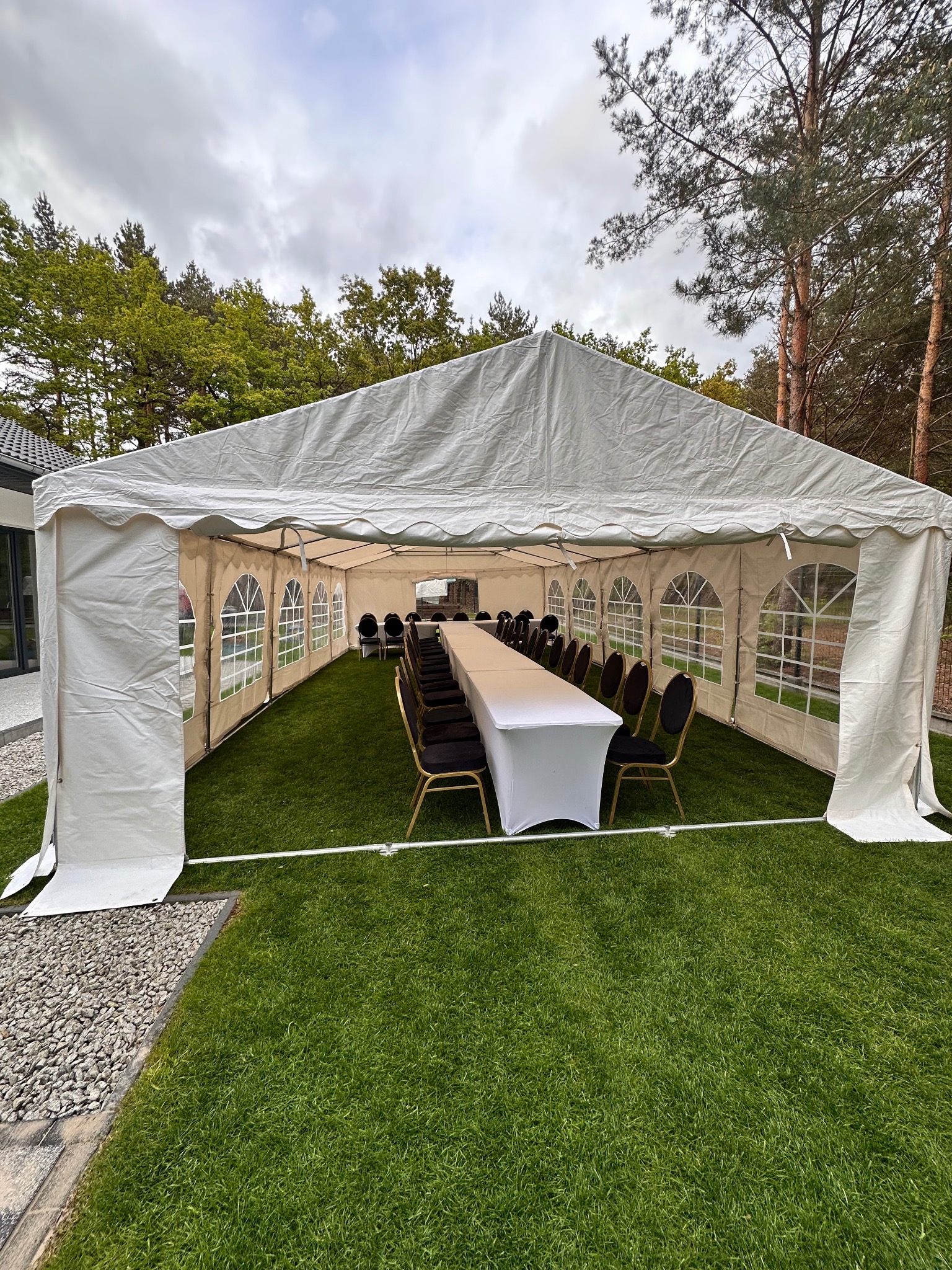 Biały namiot imprezowy na trawie, z długim stołem i krzesłami w środku. Widok z zewnątrz, w tle drzewa i pochmurne niebo. Przygotowanie do wydarzenia.