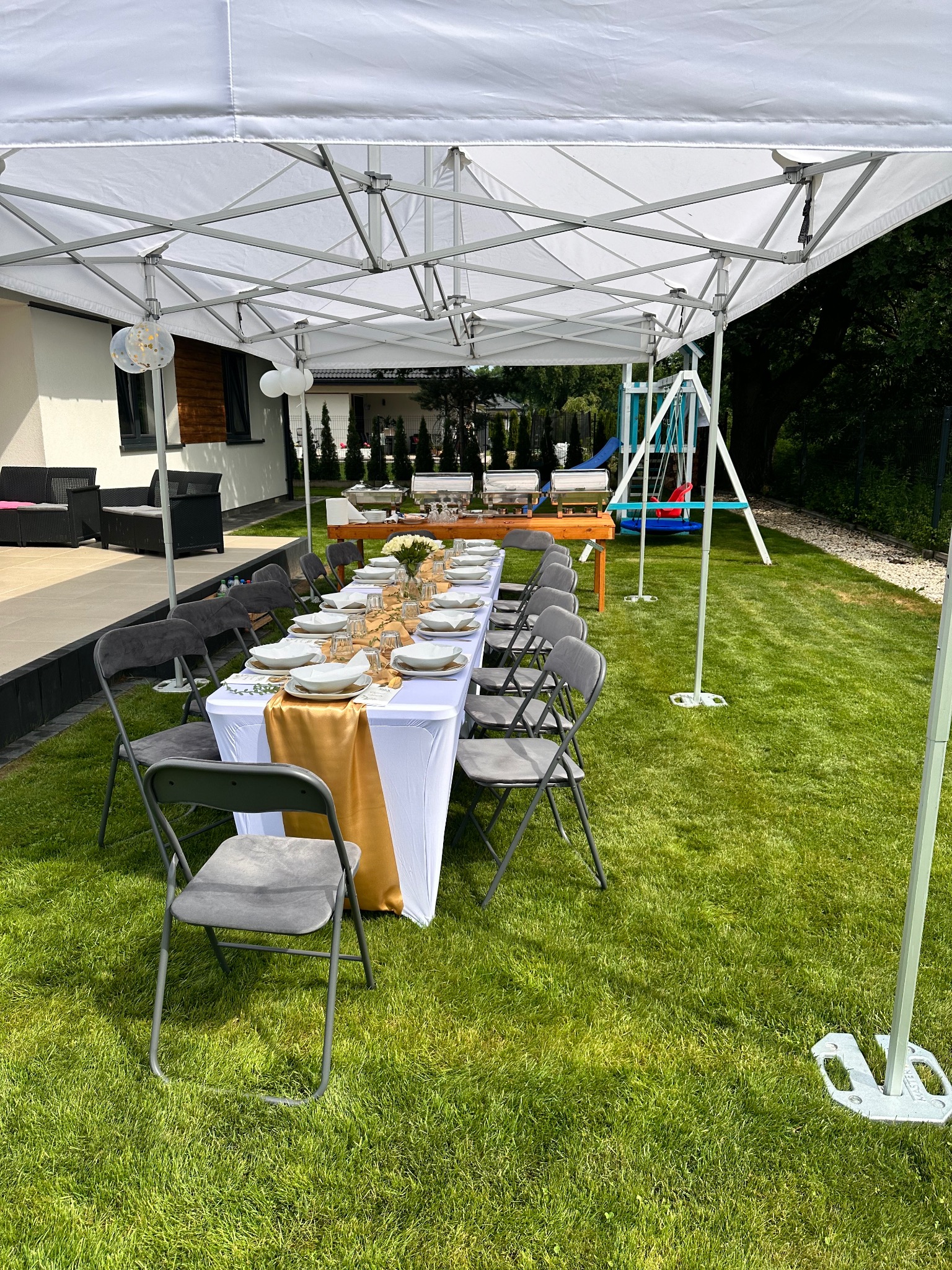 Uroczysty stół pod białym namiotem na trawie, nakryty białym obrusem ze złotym bieżnikiem, zastawiony talerzami i szklankami. W tle dom i huśtawka.