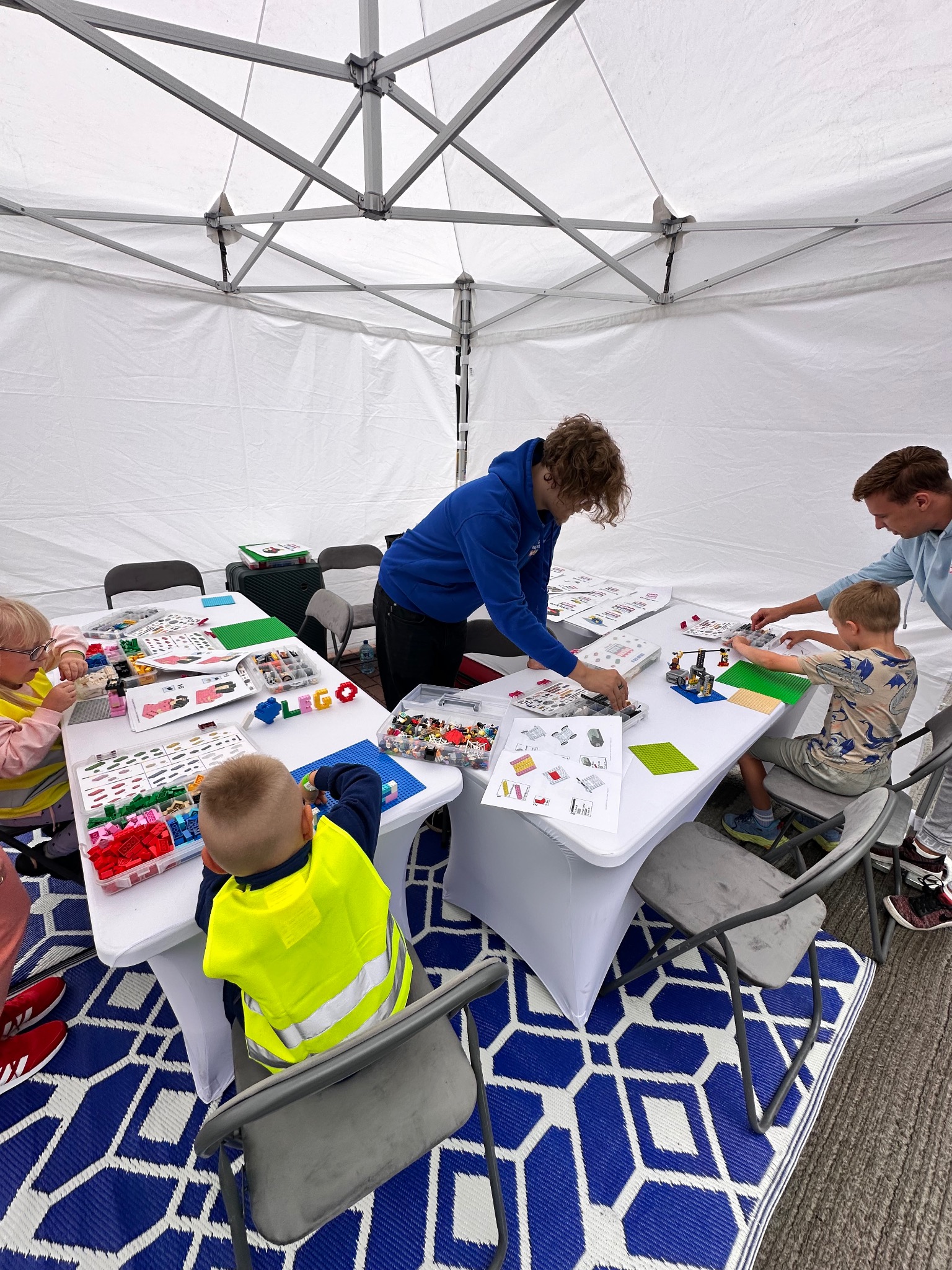 Dzieci i dorośli budują z klocków LEGO pod białym namiotem. Instrukcje, klocki rozsypane na białych stołach. Niebieski dywanik z geometrycznym wzorem.